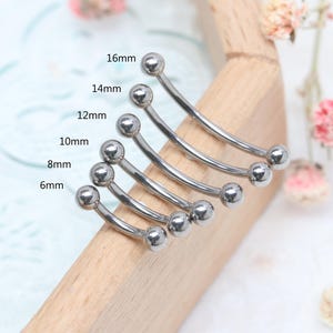 May include: A collection of curved barbell body jewelry in various sizes, ranging from 6mm to 16mm. Each barbell is silver-colored with a ball on each end. The sizes are labeled on the image.