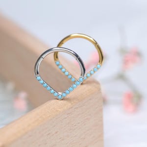 Puede incluir: Dos anillos de septum de plata y oro con un diseño en forma de lágrima y acentos de piedras preciosas azules.