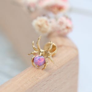 Puede incluir: Un pendiente de botón dorado con forma de araña y un centro de ópalo rosa redondo. El pendiente está colocado sobre un fondo borroso con flores rosas y blancas suaves y una superficie de madera.