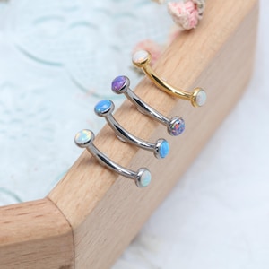 Puede incluir: Joyas curvas para cejas con piedras de ópalo en varios colores, incluyendo blanco, azul y morado. Las joyas son de metal plateado y dorado. Las piezas se exhiben sobre una superficie de madera.