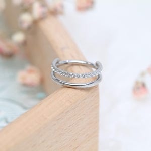 May include: A silver double-band ring, with a row of small, clear gemstones on the upper band. The ring is set against a light wooden surface, with soft, blurred floral elements in the background.