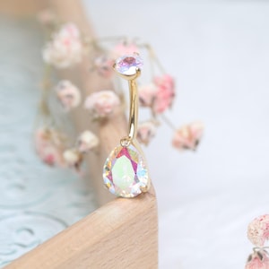 Puede incluir: Un piercing para el ombligo de color dorado con dos piedras preciosas iridiscentes, una en forma de lágrima y otra redonda. Las piedras preciosas reflejan múltiples colores. El anillo se encuentra sobre un fondo borroso de flores rosas y una superficie de madera.