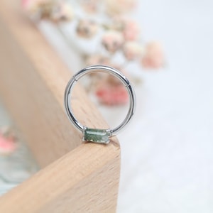 Peut inclure: Un anneau articulé de couleur argentée avec une pierre verte rectangulaire. L'anneau est posé sur une surface en bois clair avec des fleurs roses et blanches floues en arrière-plan. Bijou de piercing.