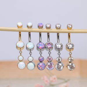 Puede incluir: Un conjunto de seis anillos de ombligo con gemas de diferentes colores. Los anillos están hechos de acero quirúrgico y tienen un encanto colgante. Las gemas son de ópalo, transparentes y rosadas.