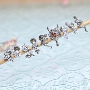 Könnte beinhalten: Eine Sammlung von silberfarbenem Körperschmuck, darunter Ohrringe und Stecker. Designs umfassen eine Schlange, eine Eidechse, eine Blume und einen Stierkopf mit einem roten Edelstein. Der Schmuck wird auf einem Holzstab vor einem weichen, verschwommenen Hintergrund präsentiert.