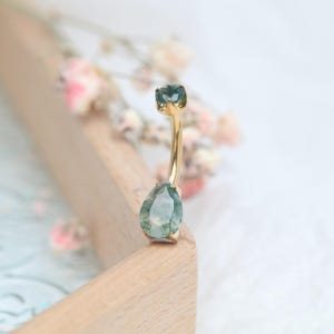 May include: A gold-colored belly button ring with two green gemstones. The top stone is square, and the bottom stone is a teardrop shape. The ring is curved and sits on a wooden surface.