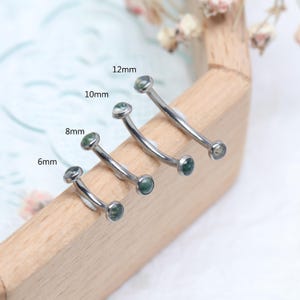 May include: Four curved eyebrow rings with green gemstone ends. Sizes are 6mm, 8mm, 10mm, and 12mm. The jewellery is displayed on a wooden surface, showcasing the different sizes available for body piercing.