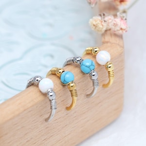 May include: Close-up of four nose rings with different colored beads. The rings have a silver and gold finish. Two rings have white beads, and two have turquoise beads. The rings are displayed on a wooden surface.