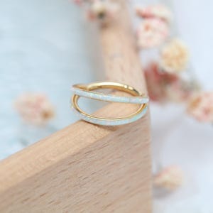 May include: A gold-colored ring with a double-band design. The bands are adorned with white opal stones. The ring is displayed on a light-colored wooden surface, with soft-focus flowers in the background.