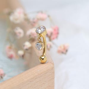 May include: A gold-colored belly button ring with two clear, round, sparkling gemstones. The top gem is fixed, while the lower gem dangles. The curved bar has a small gold ball at the bottom. The jewelry is set against a soft, blurred background.