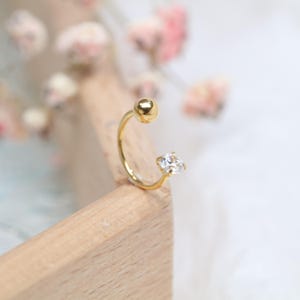 May include: A gold-coloured, open-hoop earring featuring a small, clear, square-cut gemstone at one end and a gold ball at the other. The earring is set against a light wooden surface with a blurred floral background.