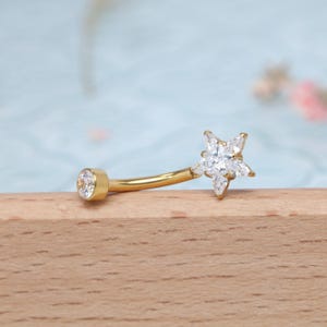 May include: A gold-colored belly button ring with a curved barbell design. One end features a clear, round gemstone, while the other end showcases a star-shaped cluster of clear gemstones. The jewelry is set against a light blue and wooden background.