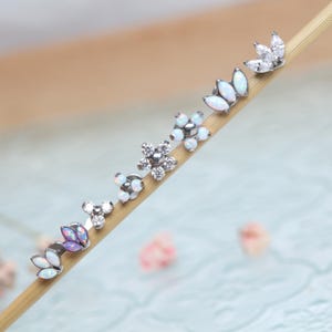 Op de afbeelding: Een verzameling zilverkleurige oorstekers met diverse bloemen- en bladvormen. De oorbellen hebben witte opaal- en heldere kristallen accenten. De oorbellen worden gepresenteerd op een houten stok tegen een zachte, vage achtergrond.