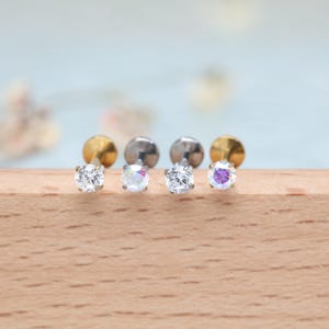 May include: Four stud earrings with round, clear gemstones. Two have gold-colored posts, and two have silver-colored posts. The gemstones are clear, iridescent, and purple. The earrings are displayed on a wooden surface.