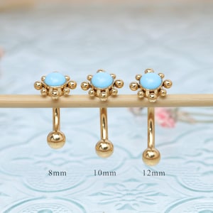 Könnte beinhalten: Drei goldfarbene Augenbrauenpiercings mit hellblauem Mittelstein und blütenförmigem Design. Die Größen sind 8 mm, 10 mm und 12 mm. Der Schmuck wird auf einem Holzstab vor einem weichen, verschwommenen Hintergrund präsentiert.