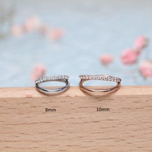 May include: Two silver and gold rings with a row of small clear stones. The ring on the left is 8mm and the ring on the right is 10mm.
