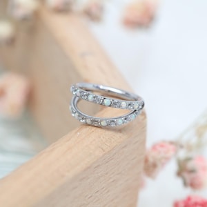 Könnte beinhalten: Ein silberner Doppelring mit kleinen, runden, weißen Opalsteinen und klaren Kristallen. Der Ring liegt auf einer hellen Holzoberfläche, mit unscharfen Blumen im Hintergrund. Der Ring ist ein Schmuckstück.