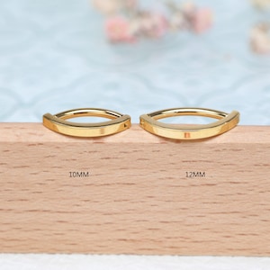 Op de afbeelding: Twee goudkleurige, gebogen sieraden. Het stuk links is gemarkeerd met "10mm" en het stuk rechts met "12mm". De sieraden worden gepresenteerd op een houten oppervlak met een lichtblauwe achtergrond.