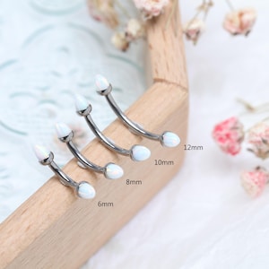 May include: Four curved eyebrow rings with white opal-like ends, in sizes 6mm, 8mm, 10mm, and 12mm. The jewelry is made of silver-colored metal and is displayed on a wooden surface.