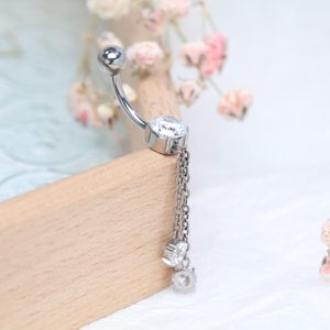 Könnte beinhalten: Ein silberfarbener Bauchnabelpiercing mit gebogener Stange. Der Schmuck hat einen großen, klaren, runden Edelstein und eine Kette mit drei kleineren Edelsteinen. Der Schmuck wird auf einer Holzoberfläche präsentiert.