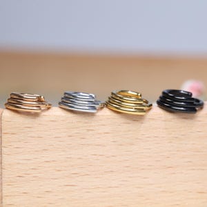 May include: Four stacked hoop earrings in different finishes: gold, silver, black, and rose gold. The earrings are displayed on a wooden surface.