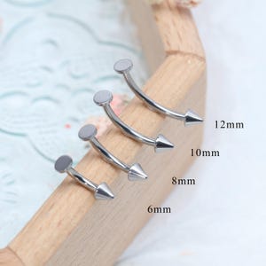 Puede incluir: Cuatro piezas de joyería de piercing para cejas curvas plateadas con extremos en forma de cono y tapas planas en forma de disco. Los tamaños son 6 mm, 8 mm, 10 mm y 12 mm. La joyería se muestra sobre una superficie de madera.