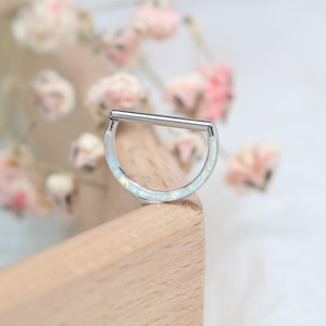 Könnte beinhalten: Ein silberfarbener, D-förmiger Körperschmuck mit einem geraden Steg oben. Der gebogene Teil ist mit einem weißen, irisierenden Material eingelegt. Der Schmuck liegt auf einer Holzoberfläche, mit verschwommenen rosa Blumen im Hintergrund.