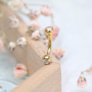 Könnte beinhalten: Ein gebogener, goldfarbener Bauchnabelring mit einem klaren, runden Edelstein an jedem Ende. Der Schmuck ist vor einem Holzblock und zarten rosa Blüten platziert.