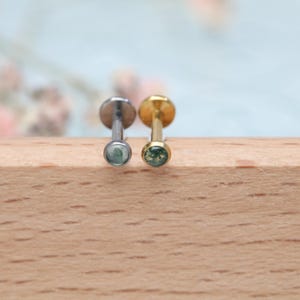 Pode incluir: Dois brincos pequenos com costas planas e uma pedra redonda verde embutida numa base de metal. Um brinco é prateado, o outro é dourado. Os brincos são exibidos em uma superfície de madeira.