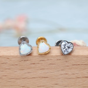 May include: Three heart-shaped stud earrings. One is silver with a white opal, another is gold with a white opal, and the third is silver with a clear, faceted stone. The earrings are displayed on a wooden surface.