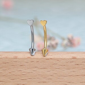 May include: Two curved eyebrow rings. One is silver-colored, the other is gold-colored. Each has a heart-shaped top and a cone-shaped bottom. The background is a soft blue with blurred floral elements, and the rings rest on a wooden surface.