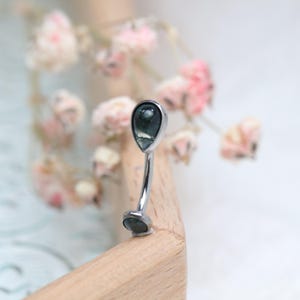 May include: A curved belly button ring with a teardrop-shaped, dark green gemstone at each end. The metal is silver-colored, and the gemstones have a marbled appearance. The background features blurred pink flowers and a wooden surface.