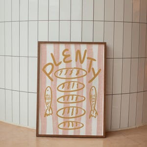 May include: A framed print with a pink and white striped background. The word "PLENTY" is written in gold script with a stack of gold bread loaves and two gold fish on either side.