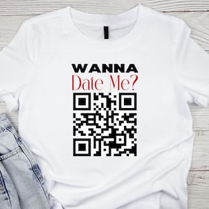 Op de afbeelding: Wit T-shirt met de tekst "Wanna Date Me?" in zwart en rood, en een zwart-witte QR-code eronder.