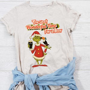 May include: A beige t-shirt with a green Grinch character holding a glass of Guinness beer and a toucan on his shoulder. The text "The Most Wonderful Time For a Beer" is printed in red and black on the shirt.