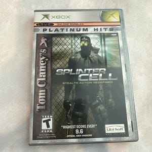 May include: A Tom Clancy's Splinter Cell video game case for the Xbox console. The cover features a character in dark tactical gear with glowing green night-vision goggles, standing before a chain-link fence. Text includes Platinum Hits, Teen ESRB rating, and Highest Score Ever 9.6.