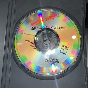 May include: A circular Sega Saturn video game sampler disc with a reflective, iridescent surface. The disc features the text 'bootleg sampler', the Sega Saturn logo, a 'Teen' ESRB rating, and 'not for resale' printed in black ink on the face.