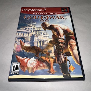 May include: A PlayStation 2 Greatest Hits video game case for God of War. The cover art features a bald warrior with chained arms holding two jagged blades in front of a classical stone building. An ESRB Mature 17+ rating is visible in the corner.