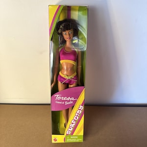 May include: A Teresa Friend of Barbie doll in a box. The doll wears a pink and yellow two-piece swimsuit. The box has the words "Surf City" and "Teresa Friend of Barbie" printed on it.