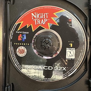 Op de afbeelding: Een Night Trap Sega CD 32X videogame disc. De schijf is zwart met rode en gele tekst, met de titel van de game en een silhouet. De schijf is gemarkeerd met "Disc 1" en is beoordeeld als "Mature".