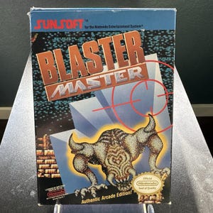 Könnte beinhalten: Eine Retro-Videospielbox für "Blaster Master" für das Nintendo Entertainment System. Die Box zeigt einen braunen und orangefarbenen Titel, eine Monsterillustration und den Text "Authentic Arcade Edition!"
