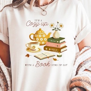May include: Cream-colored t-shirt with a graphic design featuring a teapot, teacup, books, and flowers. The text reads "It's a Cozy up with a Book kind of day."