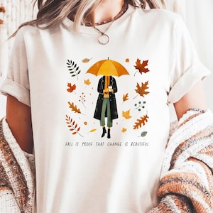 May include: Cream-colored t-shirt featuring a fall-themed illustration. The design includes a person holding an umbrella, surrounded by autumn leaves. The text "FALL IS PROOF THAT CHANGE IS BEAUTIFUL" is printed below the illustration.