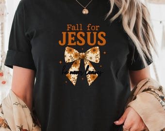 May include: Black t-shirt with the text "Fall for JESUS" in orange. A bow graphic with floral patterns is below the text, with the words "he never leaves" in script. The shirt has rolled-up sleeves.