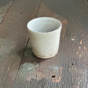 May include: A speckled ceramic cup with a cylindrical shape. The cup is off-white with small, dark brown speckles. It sits on a slightly raised base and has a smooth, rounded rim. The cup is on a weathered wooden surface.