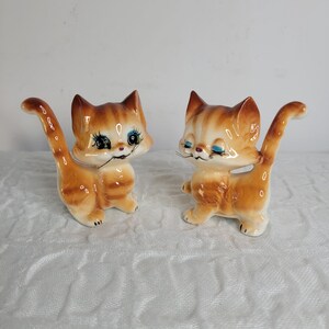 May include: Two ceramic cat ornaments. The cats are orange and white with detailed facial features, including blue eyes and black whiskers. They have arched tails and are posed in a playful stance. The ornaments are on a white surface.