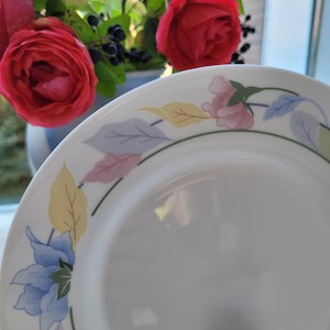Puede incluir: Plato de cena de cerámica blanca con un diseño floral. El diseño presenta flores y hojas azules, rosas, amarillas y verdes.