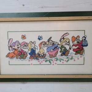 Puede incluir: Un bordado de punto de cruz enmarcado que representa a los siete enanitos de la película de Disney Blancanieves y los Siete Enanitos. Los enanitos están representados en fila, cada uno con sus características y colores distintivos. El bordado está realizado sobre un fondo blanco con detalles verdes y rosas.