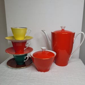 May include: A colorful tea set featuring a red teapot, sugar bowl, and stacked teacups and saucers in yellow, red, and green. The set has gold trim and a white handle and spout on the teapot.