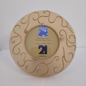 Puede incluir: Marco de fotos redondo de color crema con detalles decorativos dorados en espiral. El marco contiene una foto pequeña y presenta el texto "21" y "Garouste & Bonetti, 1995".